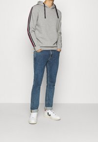 Sweat à capuche gris avec des rayures bleu marine, rouge et blanche sur les manches, cordon de serrage réglable, associé à un jean bleu et des baskets blanches.
