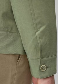 Lichtgroen shirt met een gladde textuur, voorzien van een knoopmanchet en subtiele stiksels, gecombineerd met beige broek in een casual pasvorm.