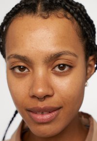 Primo piano di un volto con capelli intrecciati, occhi marroni e un anello al setto nasale. Pelle liscia con delicate lentiggini; indossa un colletto di camicia beige.