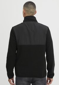 Mand med kort hår iført en sort fleecejakke med en glat panel på den øvre ryg, stående mod en ensfarvet lys baggrund.