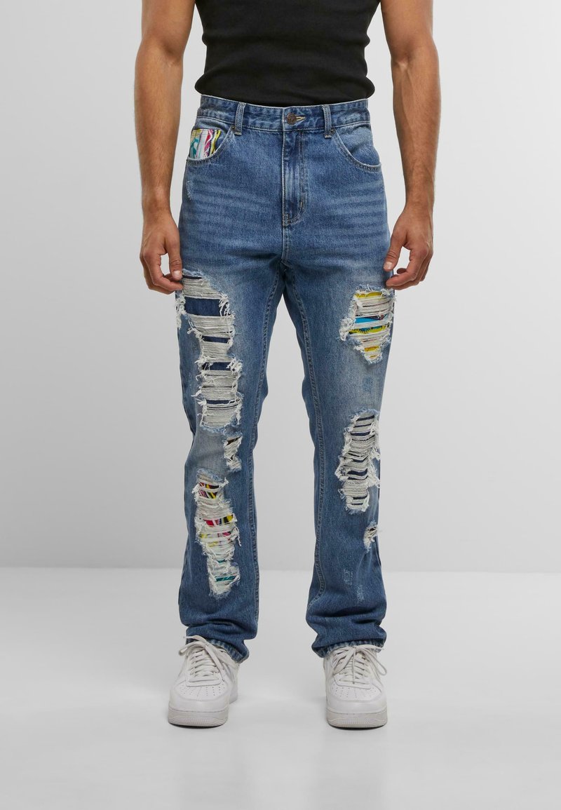 Hombre vestido con jeans azules rasgados con parches de tela coloridos debajo de las roturas, camiseta sin mangas negra y zapatillas blancas, de pie sobre un suelo blanco.