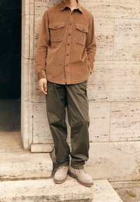 Brun fløjlsbuksedragt med to forlommer, olivengrønne cargobukser og grå støvler, stående på stenetrin mod en struktureret væg.
