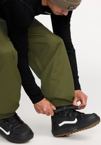 Skihose in olivgrün mit elastischen Bündchen, kombiniert mit schwarzen hohen Schuhen mit weißen Akzenten, die zur Anpassung eingestellt werden.