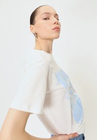 Hvid bomulds T-shirt med korte ærmer, prydet med et blåt grafisk design. T-shirten er let figursyet ved taljen og har en rund halsudskæring.
