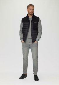 Schwarze gepolsterte Weste über einem grauen Pullover, kombiniert mit hellgrauen Jeans und schwarzen Sneakern. Die Weste verfügt über eine Brusttasche und einen Reißverschluss.