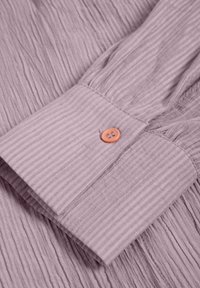 Manschetta di camicia di un leggero color lavanda realizzata in tessuto testurizzato, con strisce verticali e un accentuato bottone arancione.