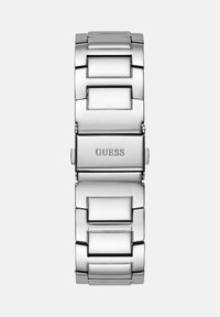 Kovinsko jekleno ura zapestnica s polirano površino, pravokotni elementi ter vgravirana oznaka "GUESS", s varnim mehanizmom zapiranja.