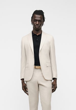 Egy világos bézs színű öltönyt viselő férfi, aki hozzáillő nadrágot, barna övet és fekete galléros inget visel, egyszínű háttér előtt áll.