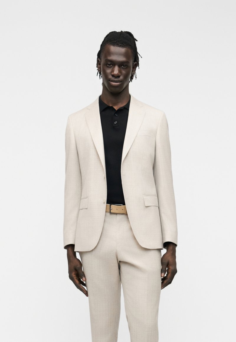 Άνδρας ντυμένος με ανοιχτό μπεζ κοστούμι και ταιριαστό παντελόνι, καφέ ζώνη και μαύρο πουκάμισο με γιακά, στέκεται μπροστά σε απλό φόντο.