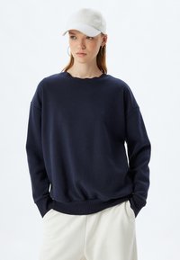 Sudadera de gran tamaño en color navy con cuello y puños de canalé, con una textura suave. Combinada con pantalones crema ajustados y una gorra blanca.