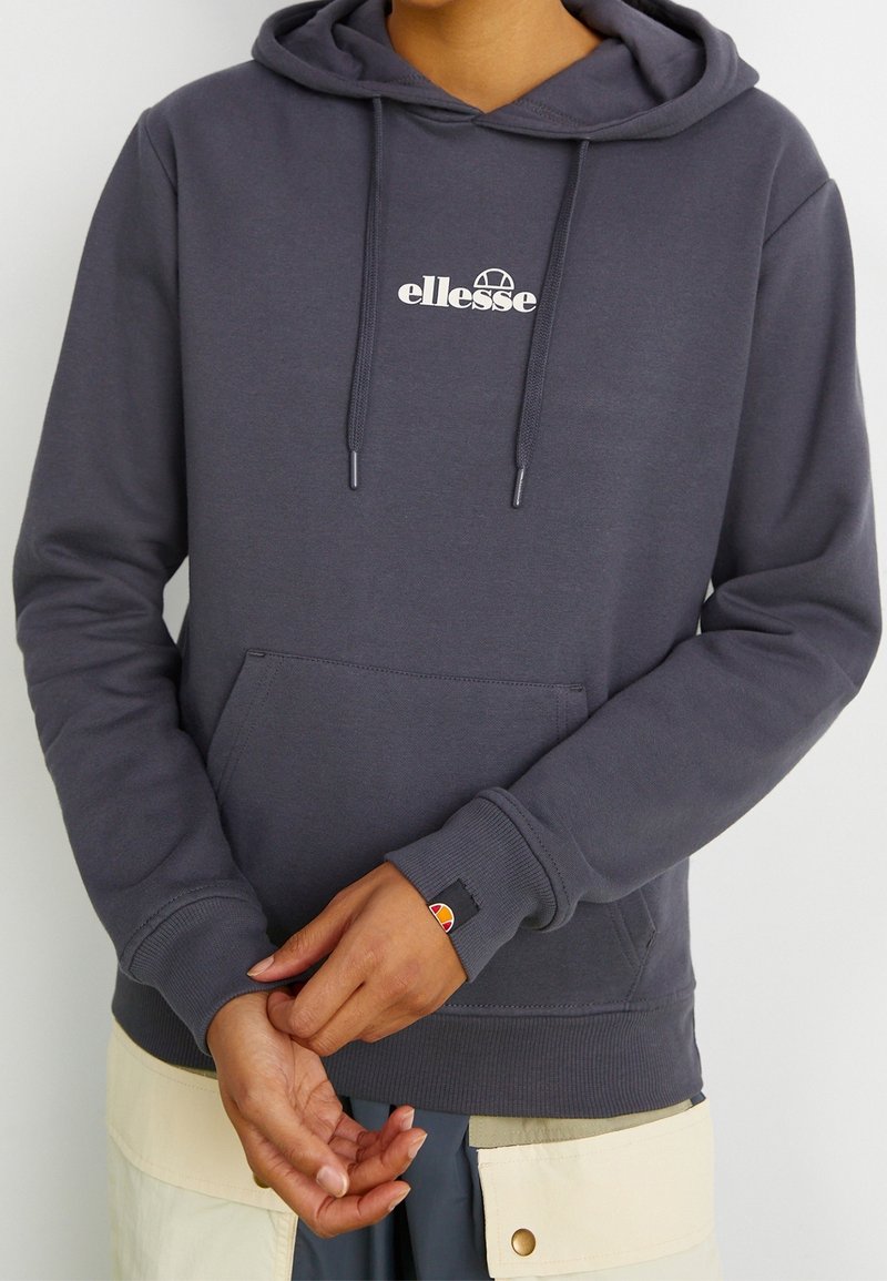 Personne portant un sweat à capuche ellesse gris foncé avec poche avant et logo, ajustant la manche, pantalon cargo beige visible à la taille.