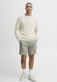 Hellbeige gestrickter Pullover mit gerippten Bündchen und Saum, kombiniert mit hellgrünen Shorts. Die Person trägt weiße Socken und graue Sneakers.