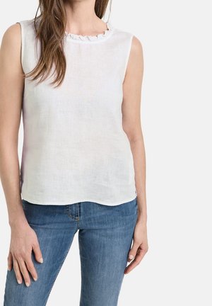 Femme portant un haut en lin blanc sans manches avec un col à volants et un jean en denim bleu, debout devant un fond clair uni.