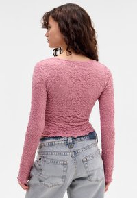 Long-sleeve top in textured pink fabric with a fitted silhouette. Features a rounded neckline and a slightly frayed hem, paired with high-waisted jeans.