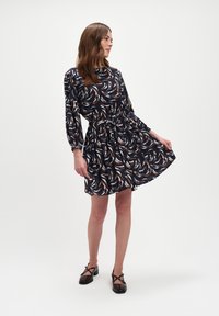 Kobieta w granatowej, wzorzystej sukience z długim rękawem trzymająca spódnicę, nosząca ciemnobrązowe, eleganckie buty Mary Jane na obcasie, na białym tle.