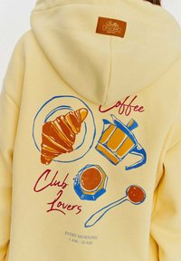 Žltá mikina s kapucňou s grafikou croissantu, kávovar a šálky. Dizajn obsahuje farebné obrysy a text: "Kávový klub milovníkov."