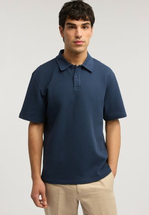 Uomo con capelli corti, ricci e scuri, che indossa una polo blu navy a maniche corte con una trama e pantaloni beige, in piedi davanti a uno sfondo semplice.