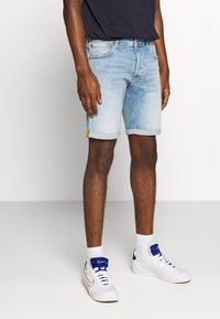 Hombre vestido con pantalones cortos de mezclilla azul claros arremangados, zapatillas Nike blancas con lengüeta azul, calcetines blancos y una camisa negra de marina, de pie sobre un suelo blanco.