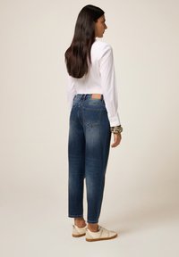 Vrouw met lang donker haar, gekleed in een witte blouse met lange mouwen, blauwe jeans, witte sneakers en een grote armband, staand met haar lichaam iets van de camera gedraaid.