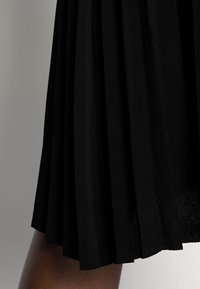 Falda negra plisada que presenta una textura suave de tela con pliegues horizontales y verticales. El dobladillo es uniforme, mostrando el diseño estructurado de la falda.