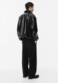 Man in een zwart leren bomberjack, losse zwarte broek en zwarte laarzen, met zijn handen in de zakken, zijwaarts gekeerd tegen een witte achtergrond.