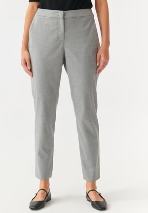Femme portant un pantalon gris clair slim avec une taille boutonnée et des ballerines Mary Jane noires, debout devant un fond uni.