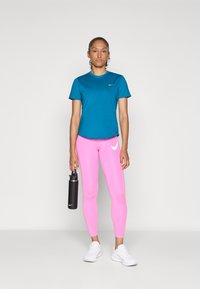 Egy nő, aki teál zöld rövid ujjú pólót és rózsaszín Nike leggingset visel, fekete vízpalackot tart a kezében, fehér sportcipő van rajta, egy sima háttér előtt.