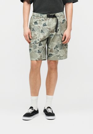 Mees, kes kannab halli kamuflaazhmustriga lühikeste püksteid musta pandlaga vööd, valgeid sokke, musti spordijalatseid ja tumehallit T-särki valge tausta vastu.