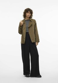 Olivgrön trenchcoat med draperad krage, stora knappar och framträdande fickor. Matchas med svarta vida byxor och en randig topp.