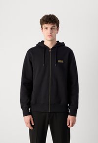 Black zip-up hoodie with a front pocket and drawstring hood. Features a small gold and black logo patch on the chest. Soft, textured fabric.