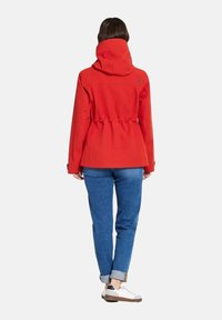 Veste imperméable rouge avec taille et capuche ajustables. Texture lisse et coupe décontractée, associée à un jean en denim bleu clair et des chaussures blanches.