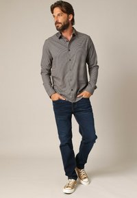 Heren overhemd met knopen in een klein ruitpatroon, grijs en zwart, met een spitskraag en lange mouwen, gecombineerd met blauwe jeans en bruine sneakers.