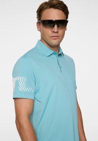 Homme portant de grandes lunettes de sport noires et un polo bleu clair avec un motif carré blanc sur la manche, debout devant un fond blanc.