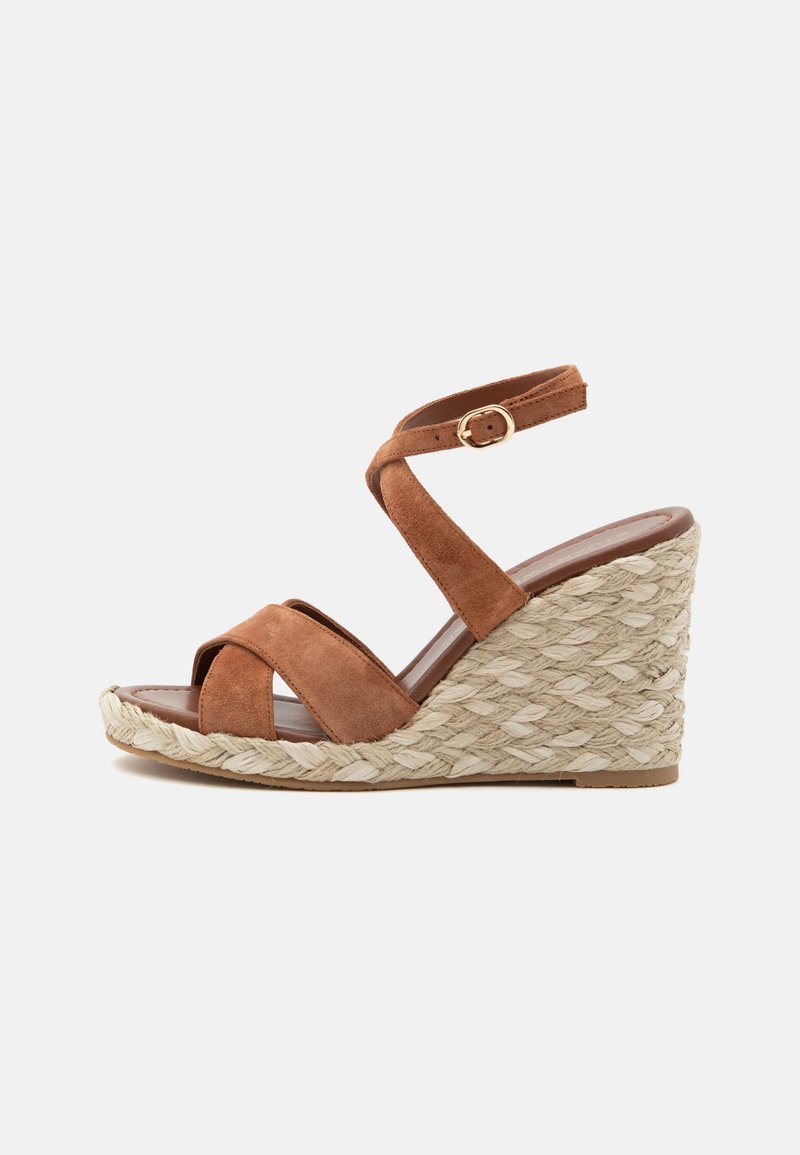 Sandale compensée en daim marron avec plateforme en jute tressée, brides croisées et boucle de cheville réglable sur fond blanc.