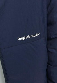 Navy plava lagana jakna s teksturiranim završetkom, sa bijelim logotipom "Originals Studio" na džepu. Zatvaranje na zatvarač.