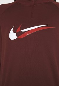 Garnetfarbener Sportpullover mit hohem Kragen, der ein großes weißes und rotes Nike-Swoosh-Logo auf der Brust trägt. Glattes, strukturiertes Material.