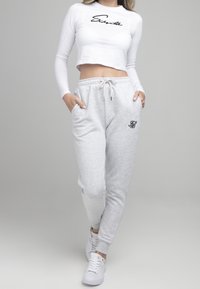 Bílý crop top s dlouhým rukávem a černým textem, spárovaný se světlými šedými tepláky s černým logem na stehně. Bílé tenisky.