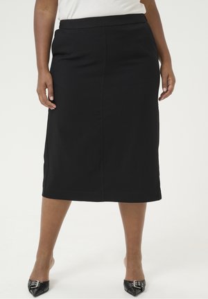 Femme portant une jupe crayon noire arrivant au genou, un haut blanc et des talons noirs à bout pointu avec des détails de boucle, debout devant un fond uni.