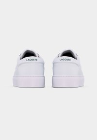 Ζευγάρι λευκών παπουτσιών Lacoste, εμφανιζόμενο από πίσω, με πράσινη ένδειξη «LACOSTE» στο πίσω μέρος της φτέρνας, σε απλό φόντο.