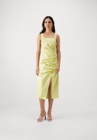 Robe midi satin jaune avec décolleté carré, détail froncé sur le côté et fente avant. Portée avec des sandales blanches à lanières. Texture lisse.