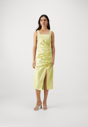 Gul satin midi-kjole med firkantet utringning, sammenrullende detaljer på siden og splitt foran. Kombinert med strappy hvite hæler. Glatt tekstur.