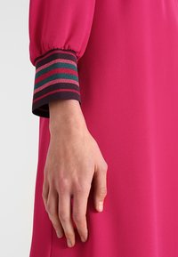 Vestido fucsia con una textura suave; presenta un puño a rayas en verde, rosa y burdeos. Una mano descansa sobre el vestido.