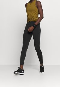 Osoba oblečená v hořčičkovém bezrukávovém topu, černých legínách, černém náramku Nike a černých teniskách Nike, stojící na prázdném bílém pozadí.