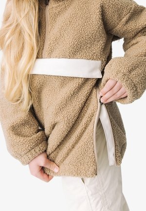 Osoba rozpinająca boczny zamek beżowej kurtki z polaru sherpa, o długich jasnych włosach i jasnych spodniach, na białym tle.