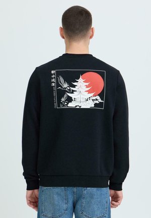 Mees, kes kannab musta dressipluusi, millel on valge Jaapani pagood, kaks kraanat, punane päike ja tagaküljel kiri "Vaikne vesi voolab sügavalt".