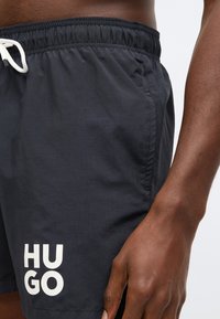 Juodi maudymosi šortai su elastinga juosmens juosta ir raišteliu, kairėje kojų pusėje yra baltas „HUGO“ logotipas. Lygus, lengvas audinys.