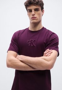 Maroon cotton t-shirt with a round neck and short sleeves, featuring an embossed "CK" logo on the chest. Smooth texture, minimal design.