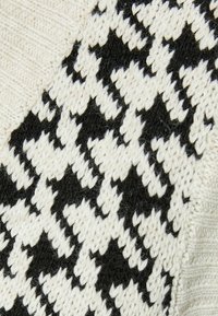 Neulepusero, jossa on musta ja kerma houndstooth-kuviointi, yhdistettynä teksturoituun kankaaseen ja sileisiin, yksivärisiin osiin.