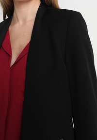Svart skräddarsydd kavaj med spetsig krage och rak passform, bärs över en djupröd blus. Har slät tyg och subtila sömnadsdetaljer.