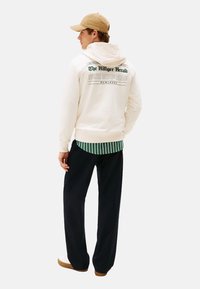 Hombre con una gorra beige, sudadera blanca con el texto "The Hilfiger Herald", camiseta a rayas verdes, pantalones negros y zapatillas blancas, de pie de espaldas.